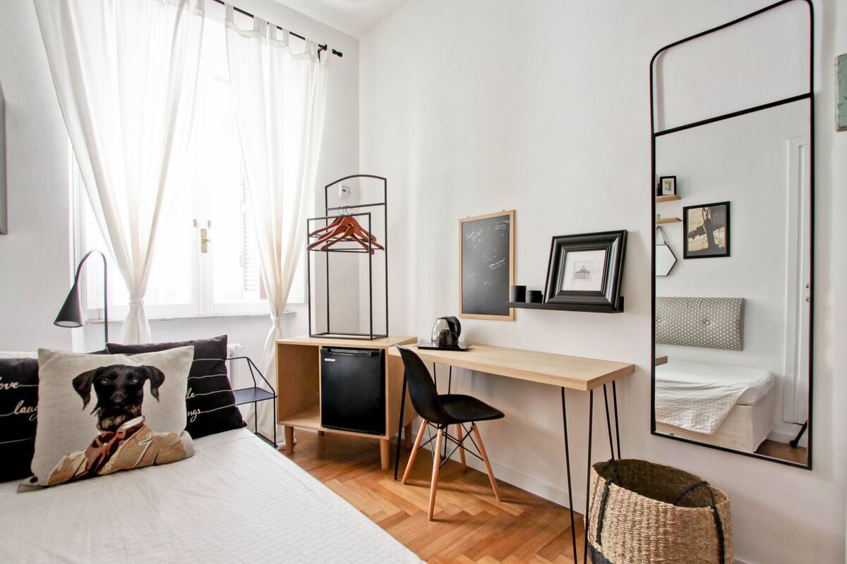 A bedroom with a bed, a desk, a chair, a small refrigerator, a mirror, and a window with curtains.