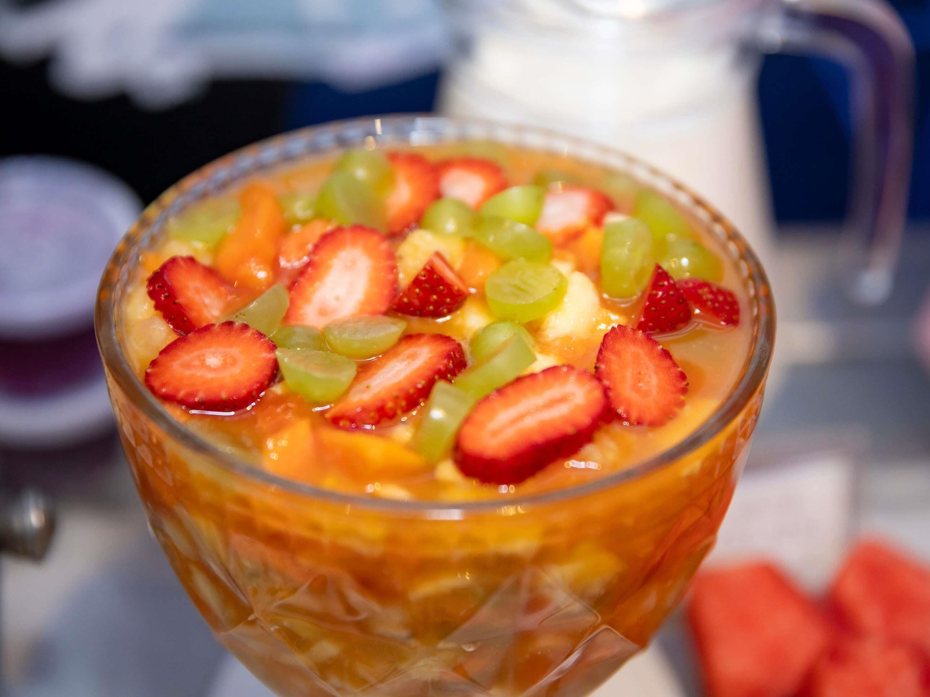 Un saladier en verre rempli d’une salade de fruits, notamment de fraises et de raisins, est posé sur une table.