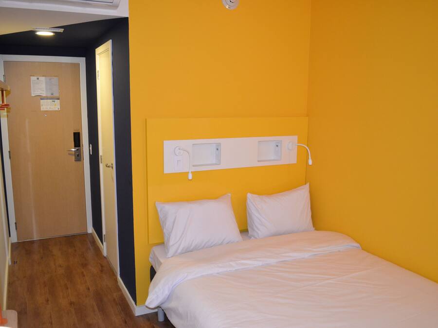 Una habitación de hotel con una cama, piso de madera, pared amarilla y una puerta con un tablón de anuncios.