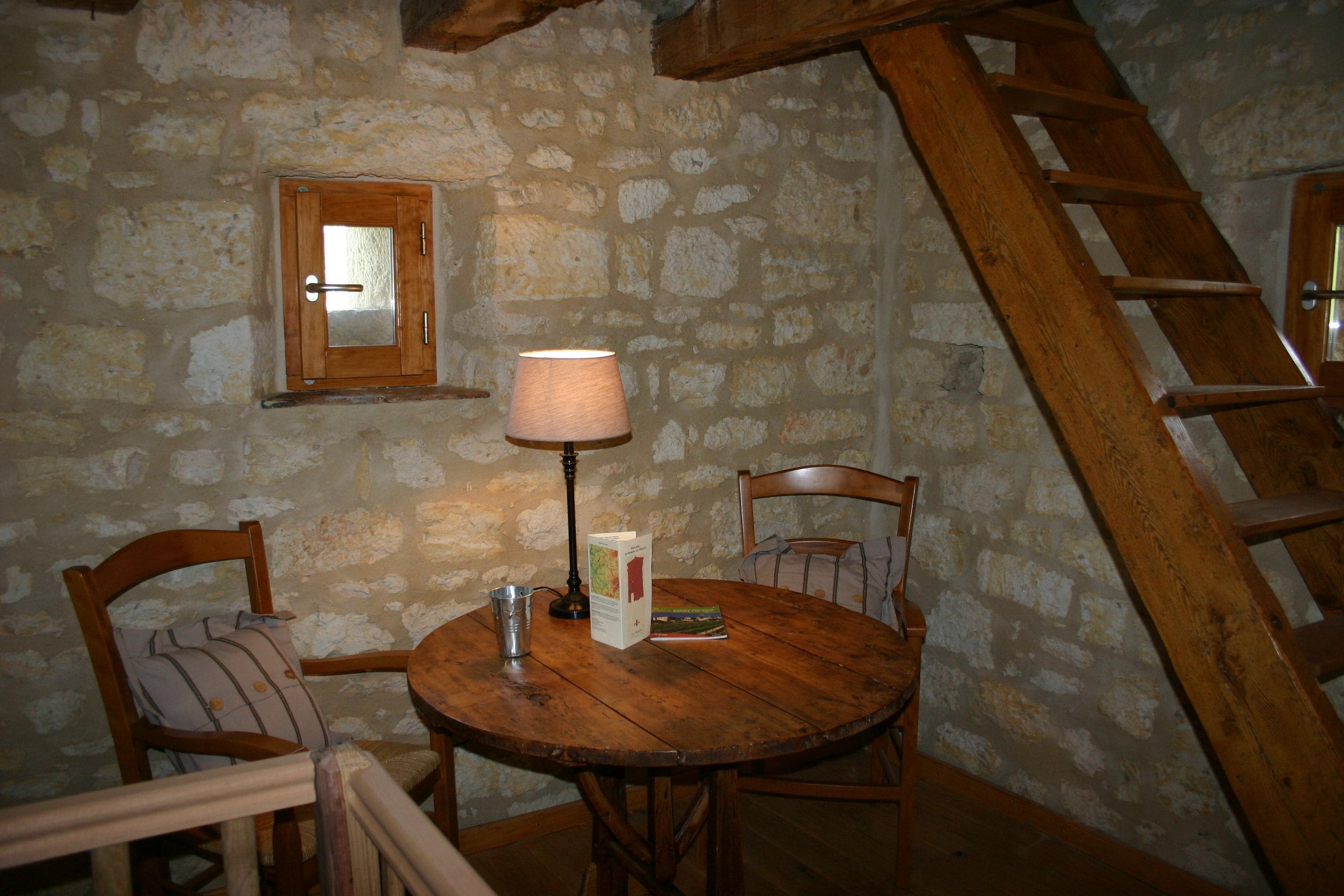 Una habitación rústica con paredes de piedra, una mesa de madera, sillas, una lámpara y una escalera.