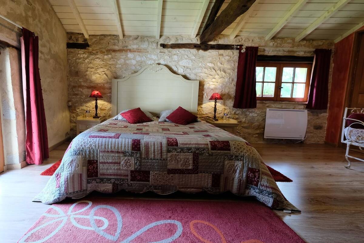 Une chambre avec un mur en pierre, un plafond en bois, un grand lit recouvert d’une courtepointe, deux lampes de chevet et un radiateur.