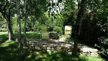 Un mur de pierre, deux chaises vertes et une table dans un coin de jardin à l’ombre.