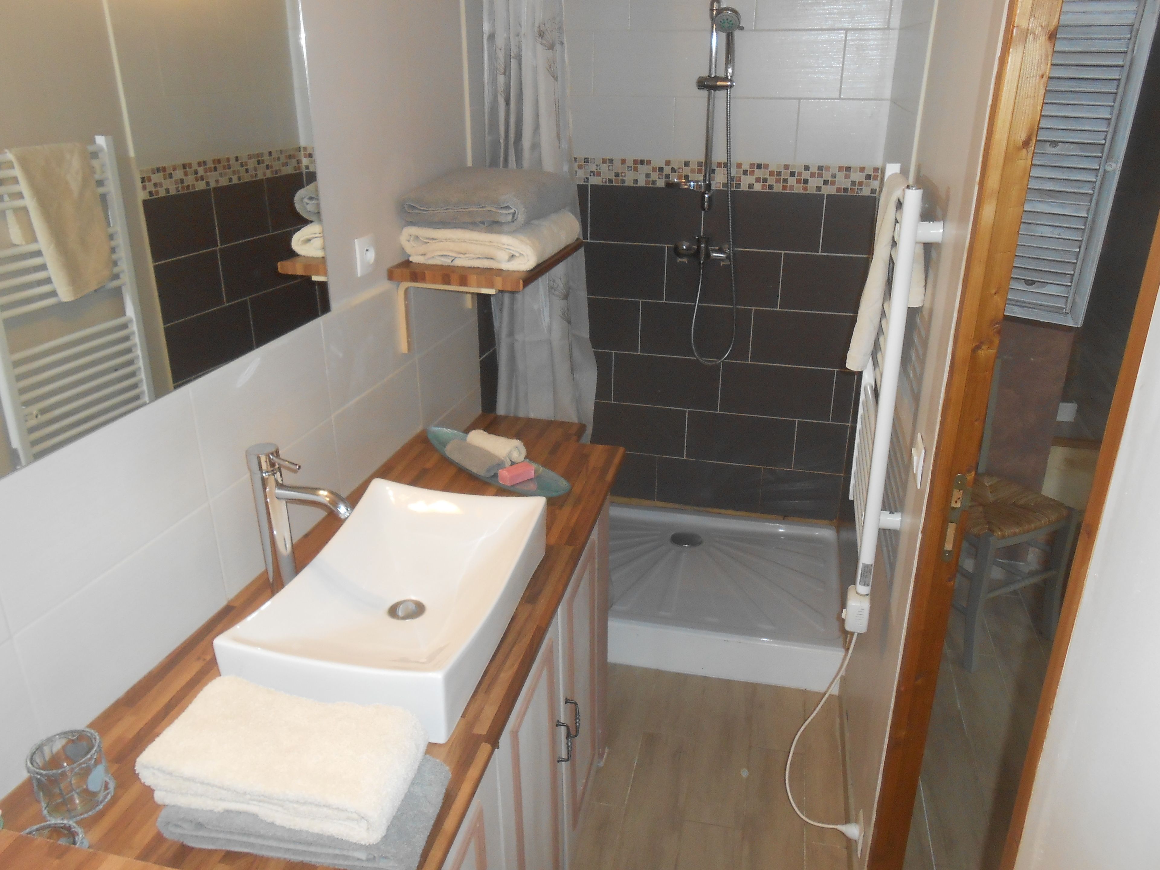 A bathroom with a sink, mirror, shower, and a small wooden shelf with towels.