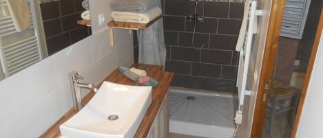 A bathroom with a sink, mirror, shower, and a small wooden shelf with towels.