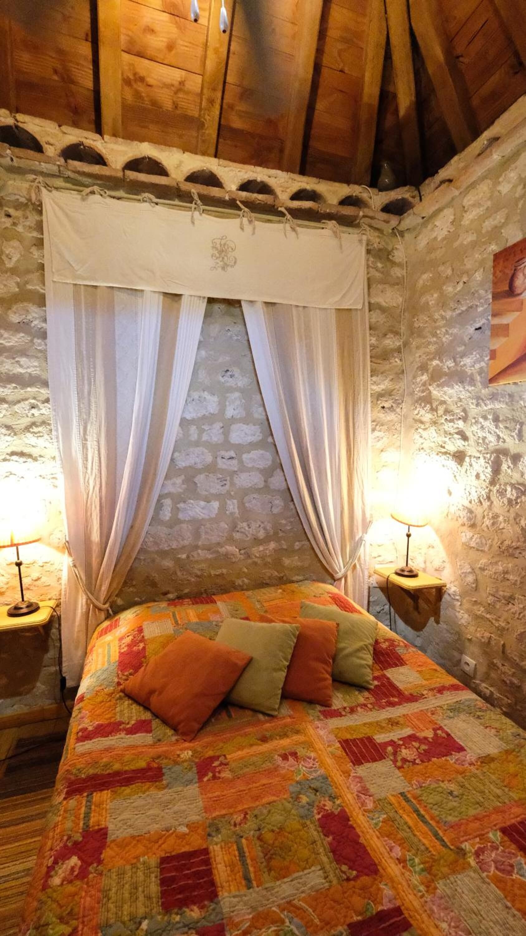 Un dormitorio rústico con techo de madera, pared de piedra, cama con colcha de colores y dos lámparas de noche.