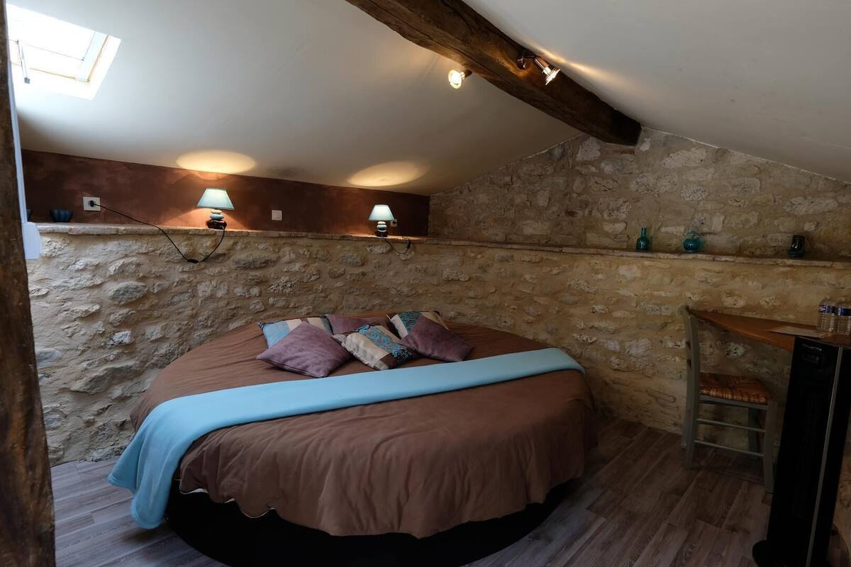 Une chambre avec un mur en pierre, un lit agrémenté de plusieurs coussins, une petite étagère avec des objets décoratifs et un plafond avec une poutre en bois.