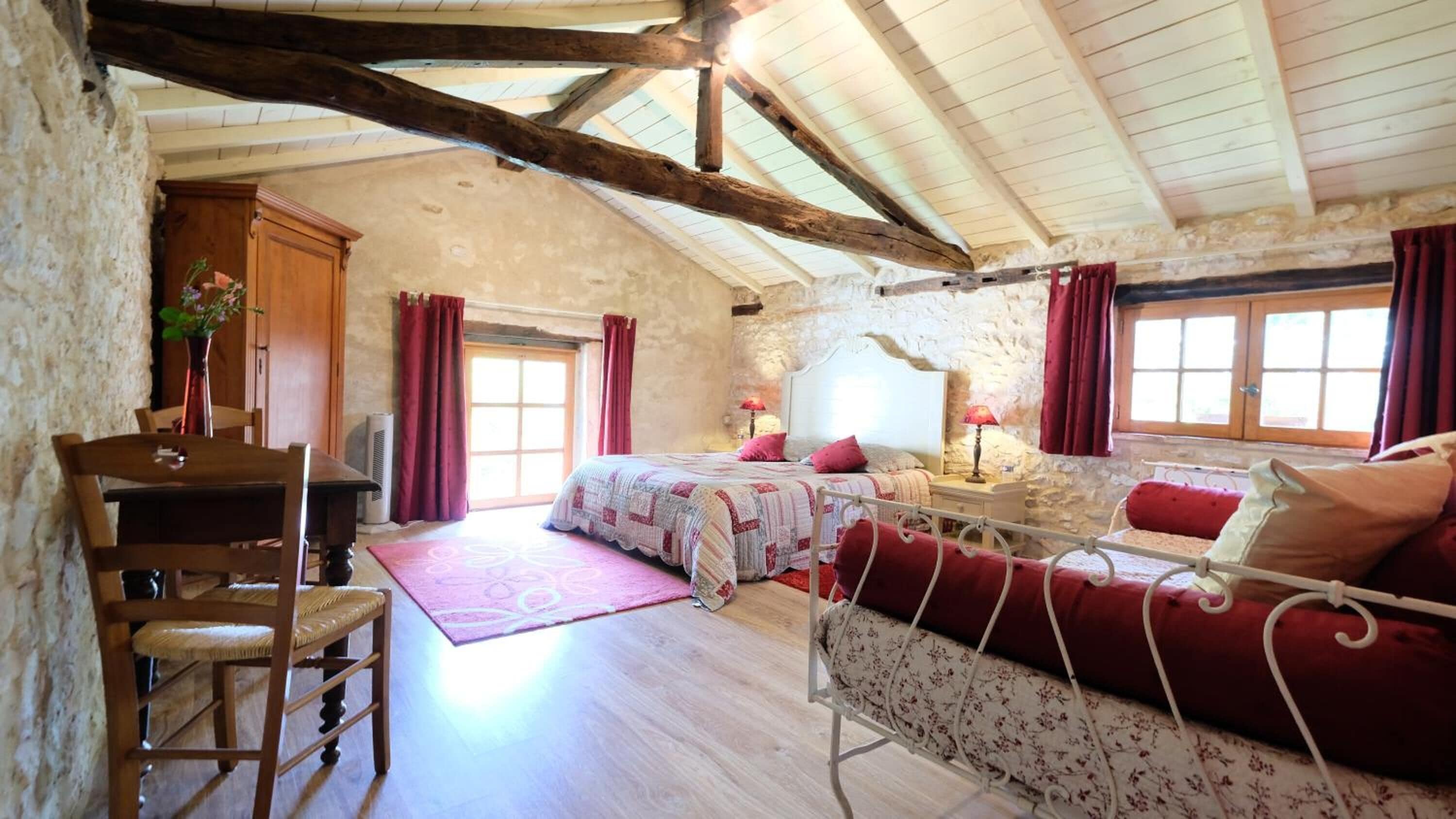 Une chambre à coucher rustique avec un plafond en bois, un lit avec une literie rouge, une chaise et un bureau agrémenté d’un vase de fleurs.