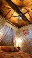 A bedroom with a wooden ceiling, a bed with colorful bedding, a nightstand with a lamp, and a painting on the wall.