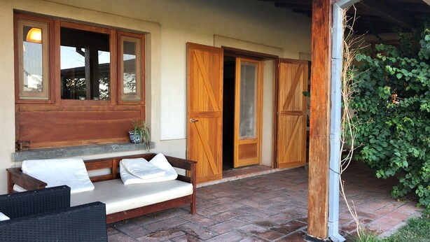 A covered outdoor seating area with a bench and a table, wooden doors, and a brick floor.