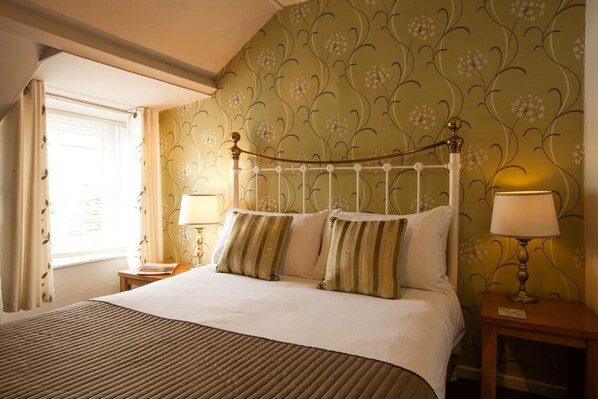A bedroom with a bed, two bedside tables, a window with curtains, and a floral wallpaper pattern.