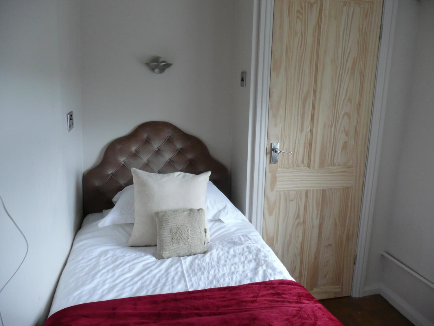 Un dormitorio con cabecera de madera, colcha blanca, manta roja y puerta de madera.