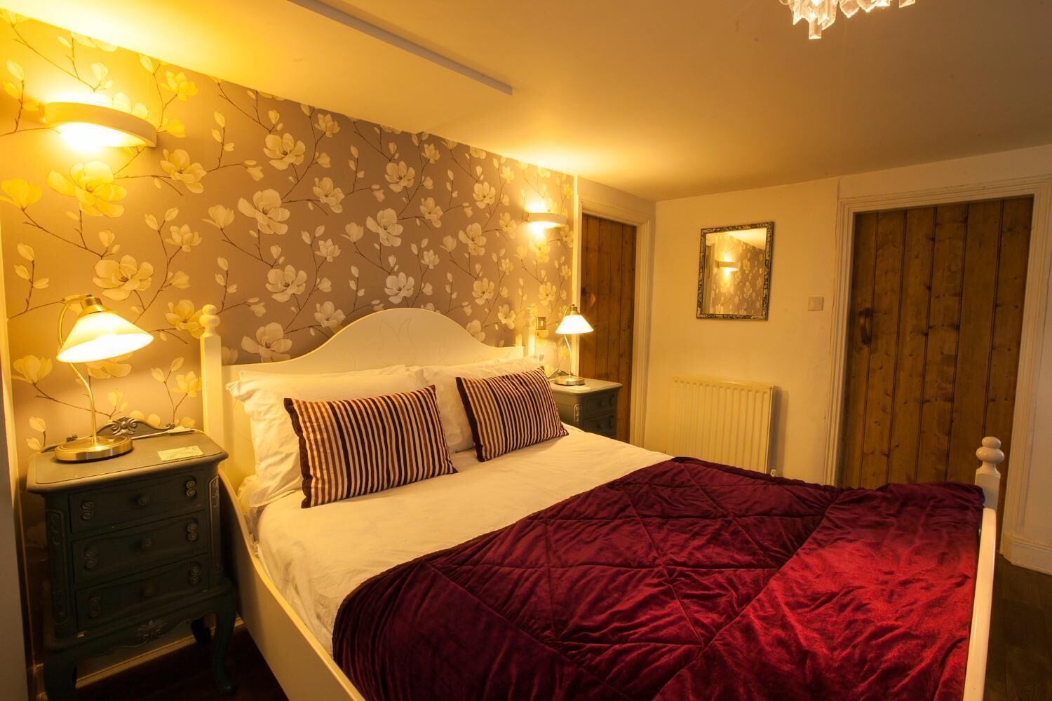 Ein Schlafzimmer mit einem großen Bett, zwei Nachttischlampen, blumigem Wandpapier, einer Holztür und einem Spiegel an der Wand.