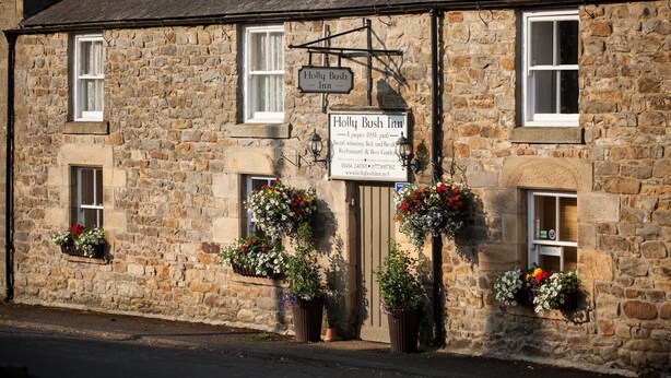 A stone building with a sign for the Holly Bush Inn, featuring flower baskets and a signboard advertising its services. - Holly Bush Inn (Hexham)