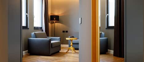 Una habitación de hotel moderna con piso de madera en patrón de espiga, dos sofás grises, una mesita redonda y una lámpara de pie.