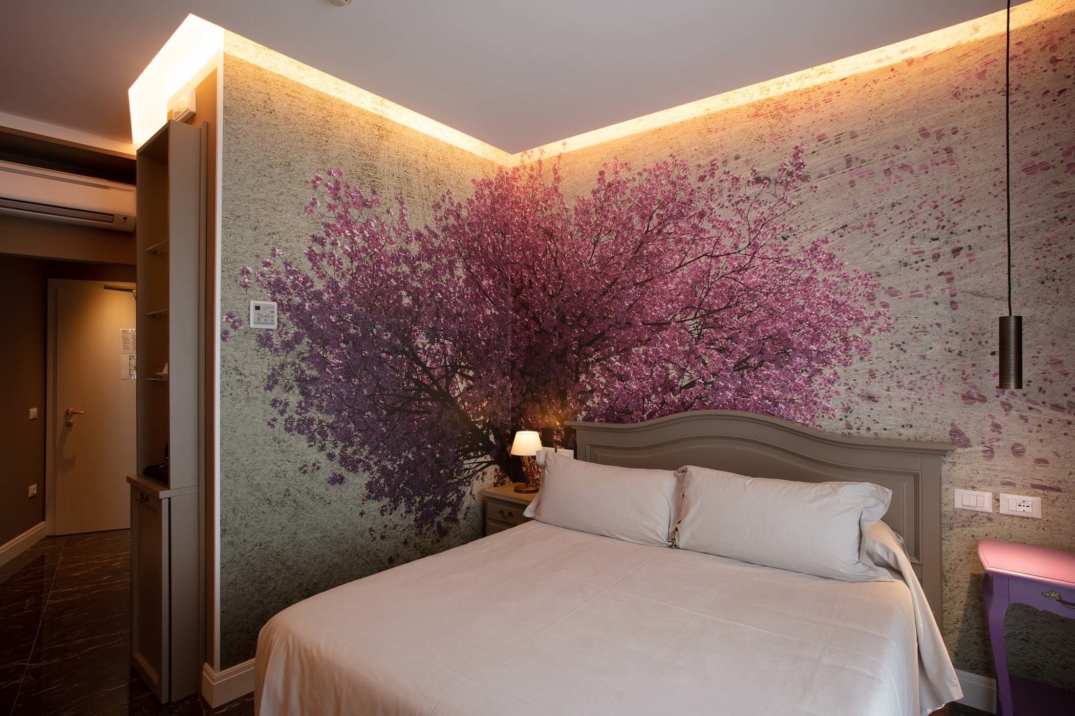 Habitación de hotel con cama, mesita de noche, lámpara y un mural en la pared con un árbol.
