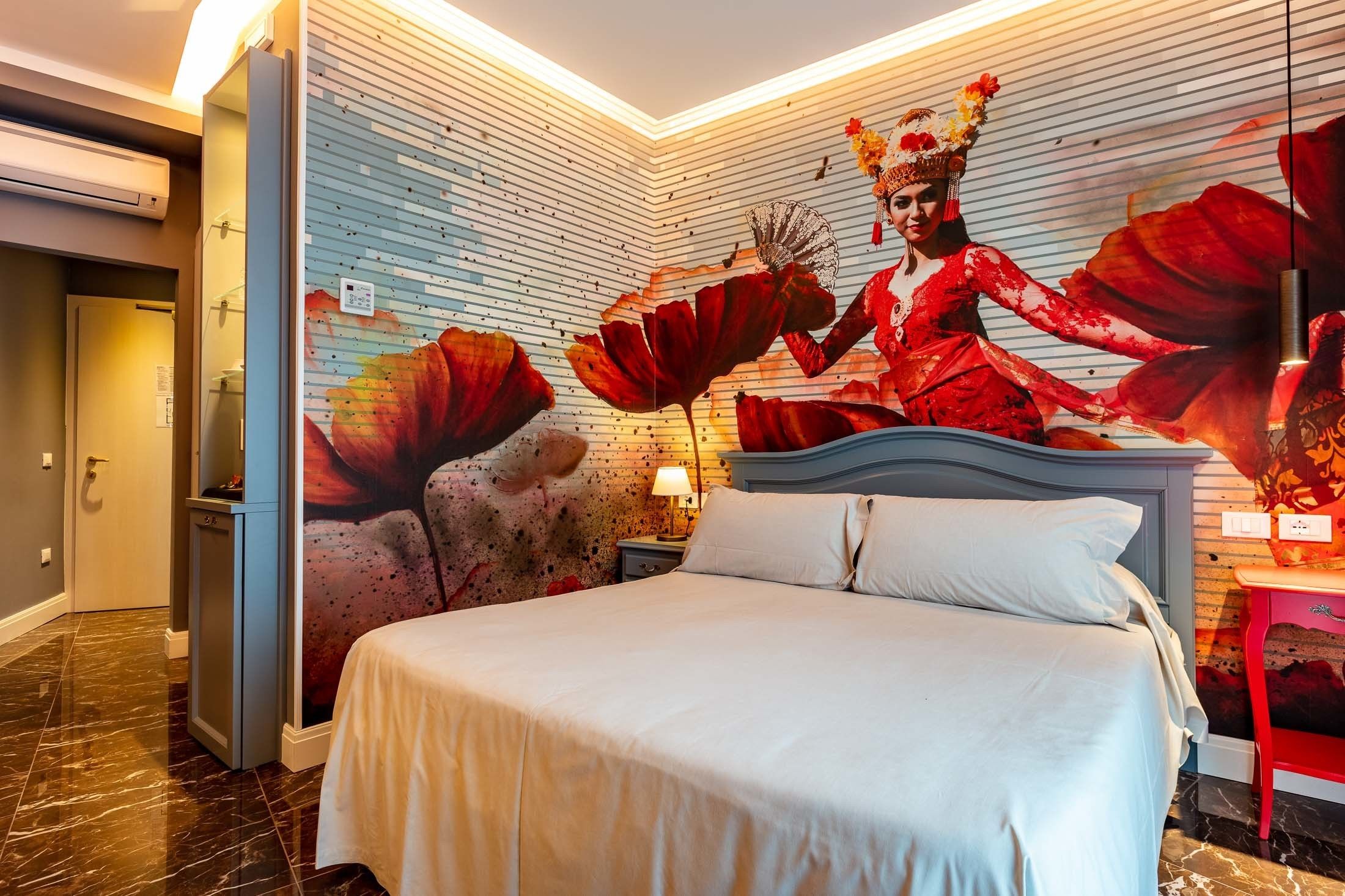 A hotel room with a large bed, a nightstand with a lamp, a wall mural, and a red side table.