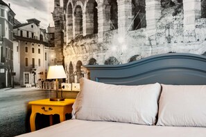Un lit avec du linge de lit blanc et une tête de lit bleue, une table de chevet jaune avec une lampe, et une fresque représentant des ruines antiques en arrière-plan.