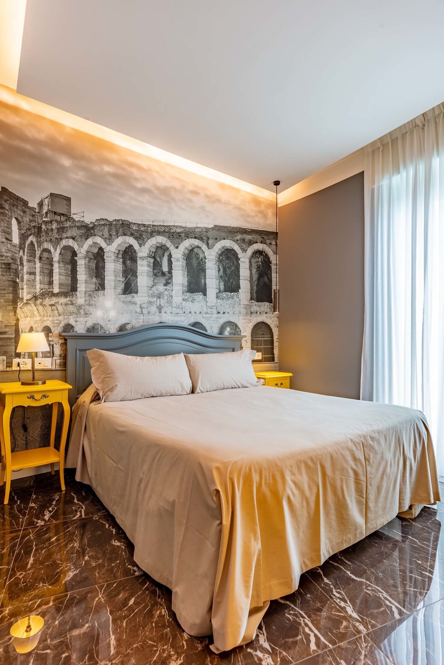 Quarto de hotel com cama, mesa de cabeceira, luminária, espelho e um mural na parede.