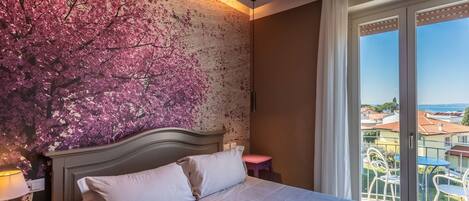 Une chambre avec un lit, une table de chevet, un tabouret rose, une fenêtre donnant sur des immeubles et un mur orné d’une fresque représentant des fleurs de cerisier.