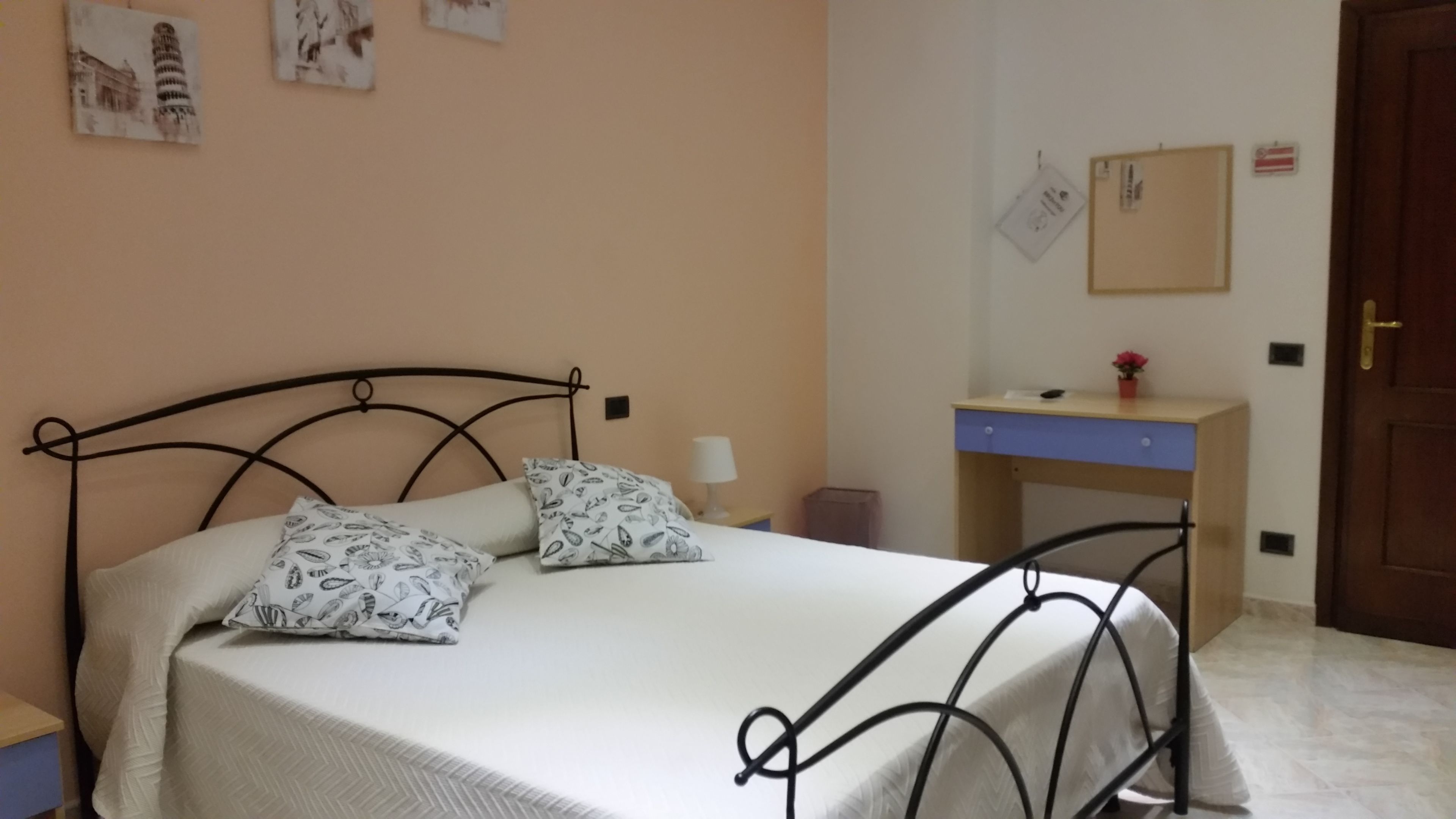 A neatly made bed with patterned pillows, a bedside table with a lamp, a desk with a computer, and a door with a lock.