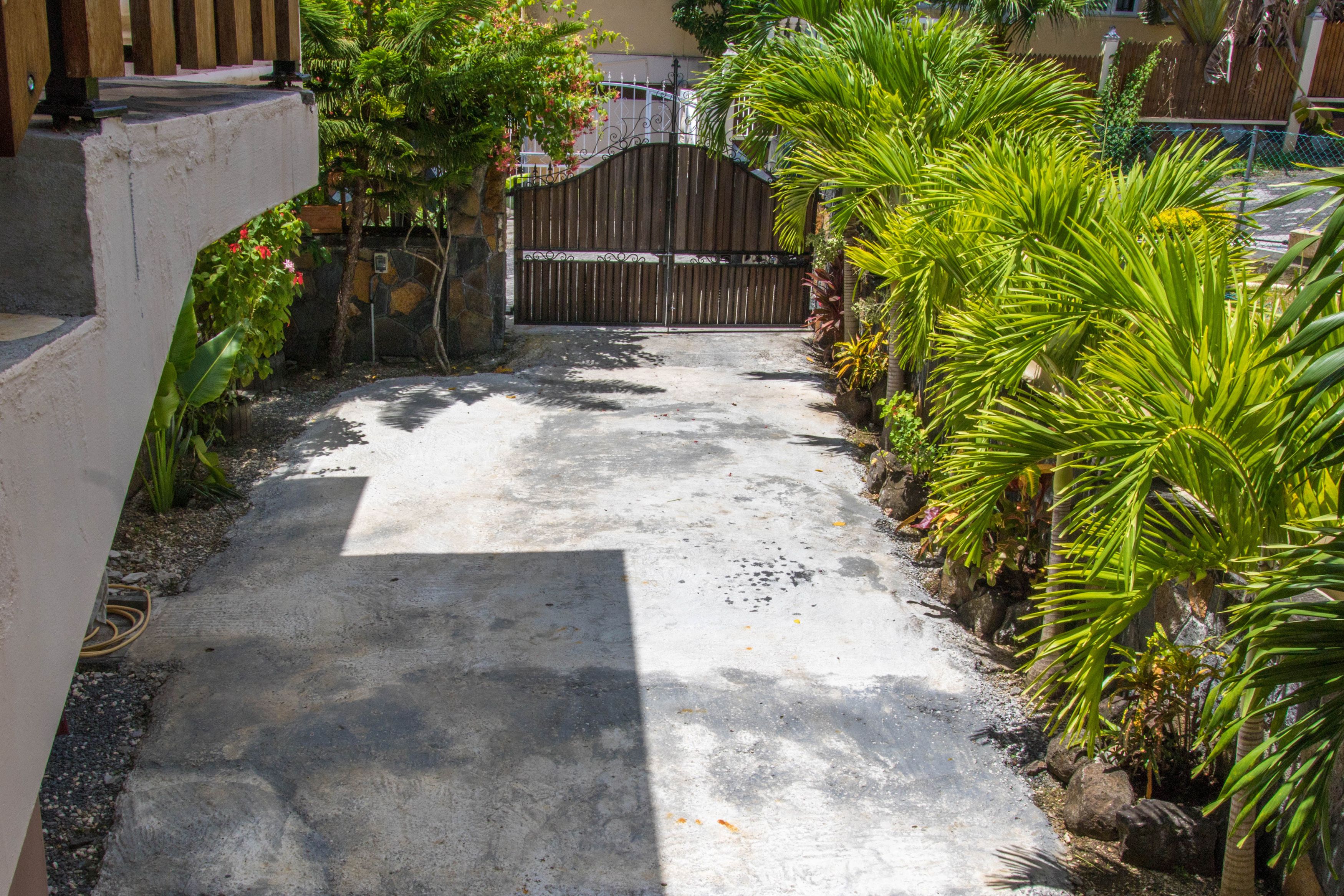 A concrete pathway leading to a gated entrance, surrounded by lush greenery and a stone wall.