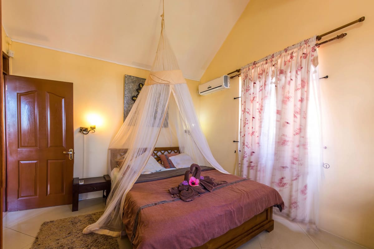 Un dormitorio con una cama de madera, mosquitera, cortina floral, puerta de madera y aire acondicionado instalado en la pared.