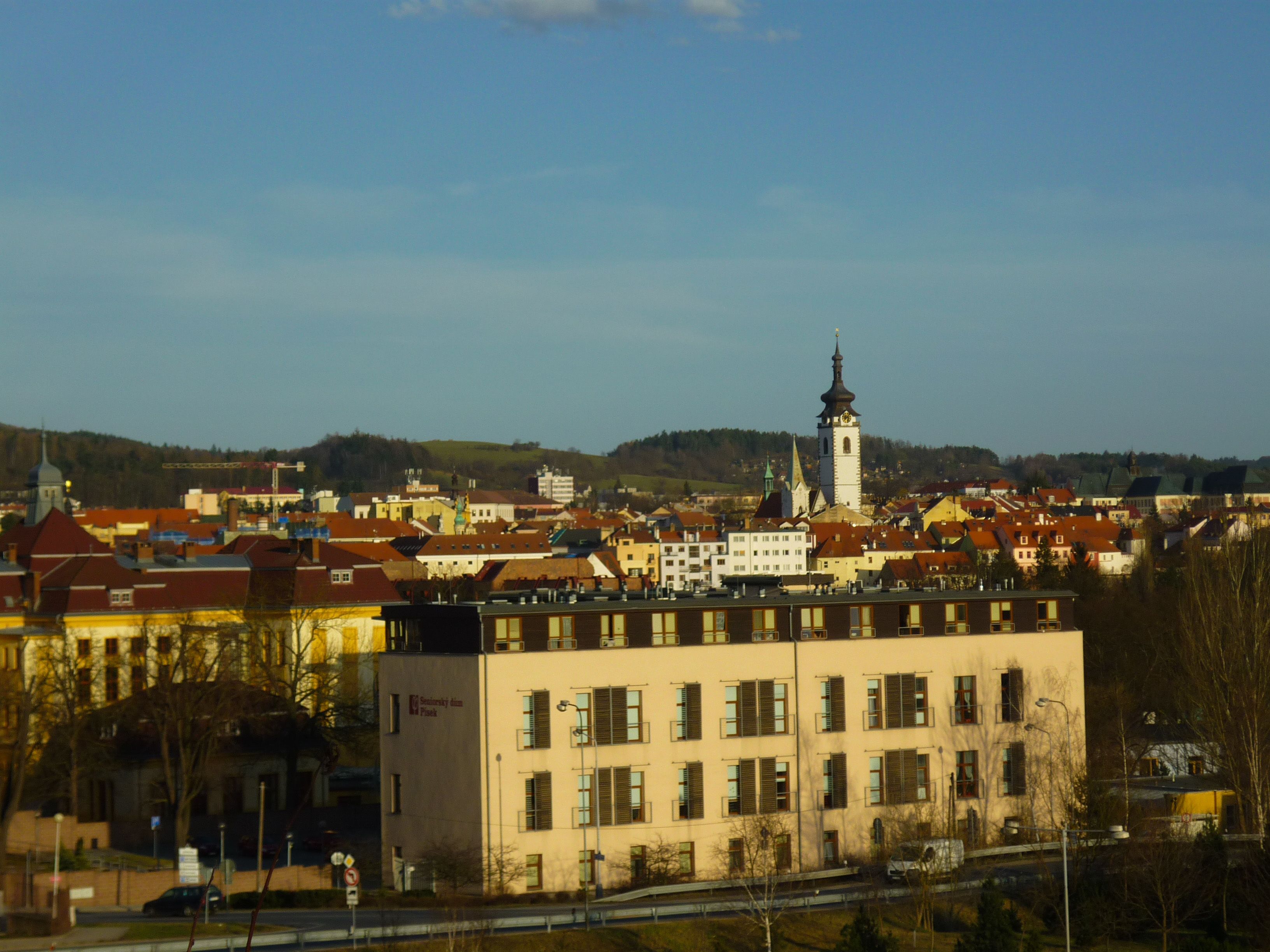 A town with a prominent church tower, a mix of residential and commercial buildings, and a flat terrain with hills in the background.