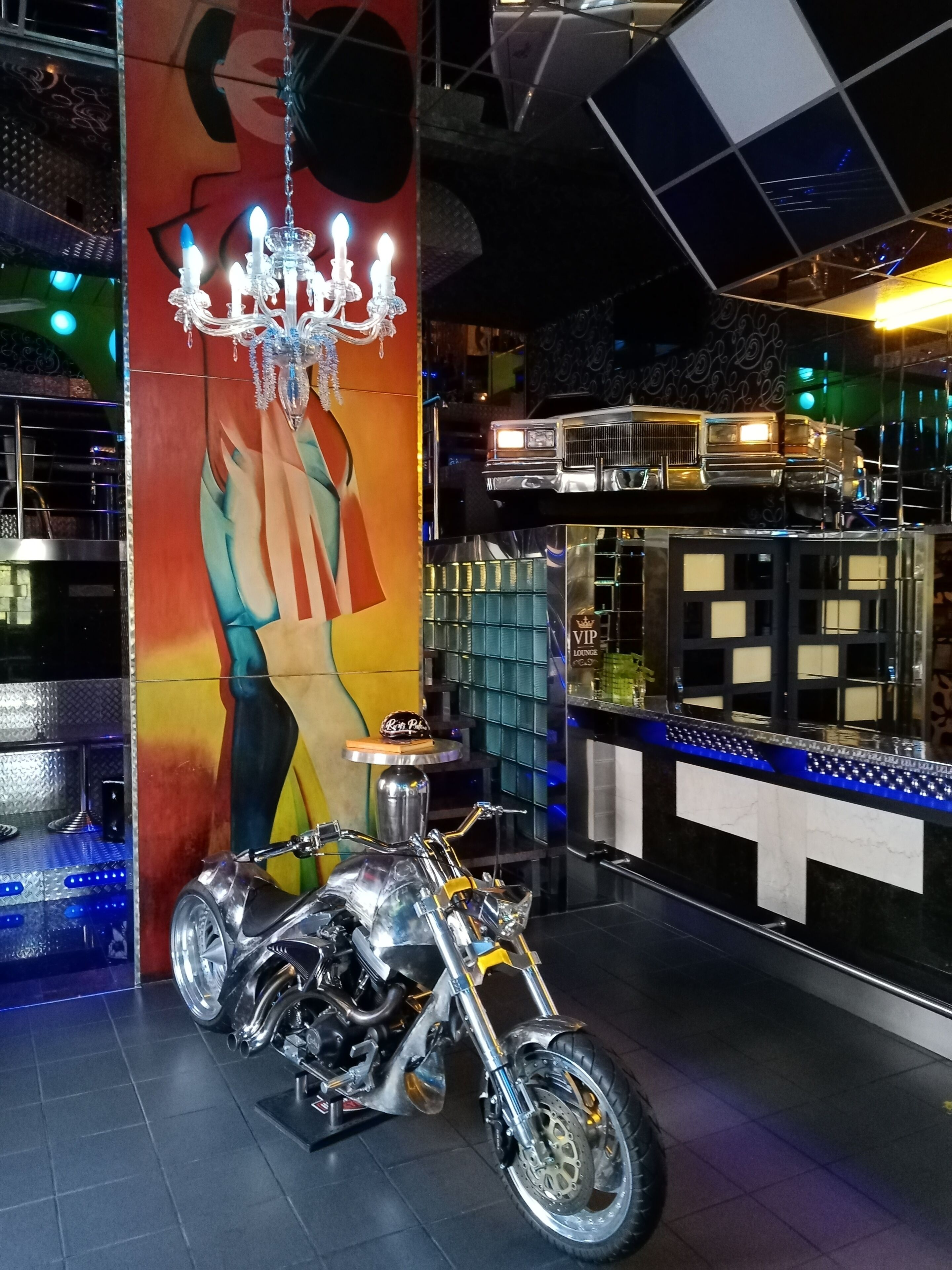 A motorcycle displayed in a room with a colorful mural, a chandelier, and a bar area.