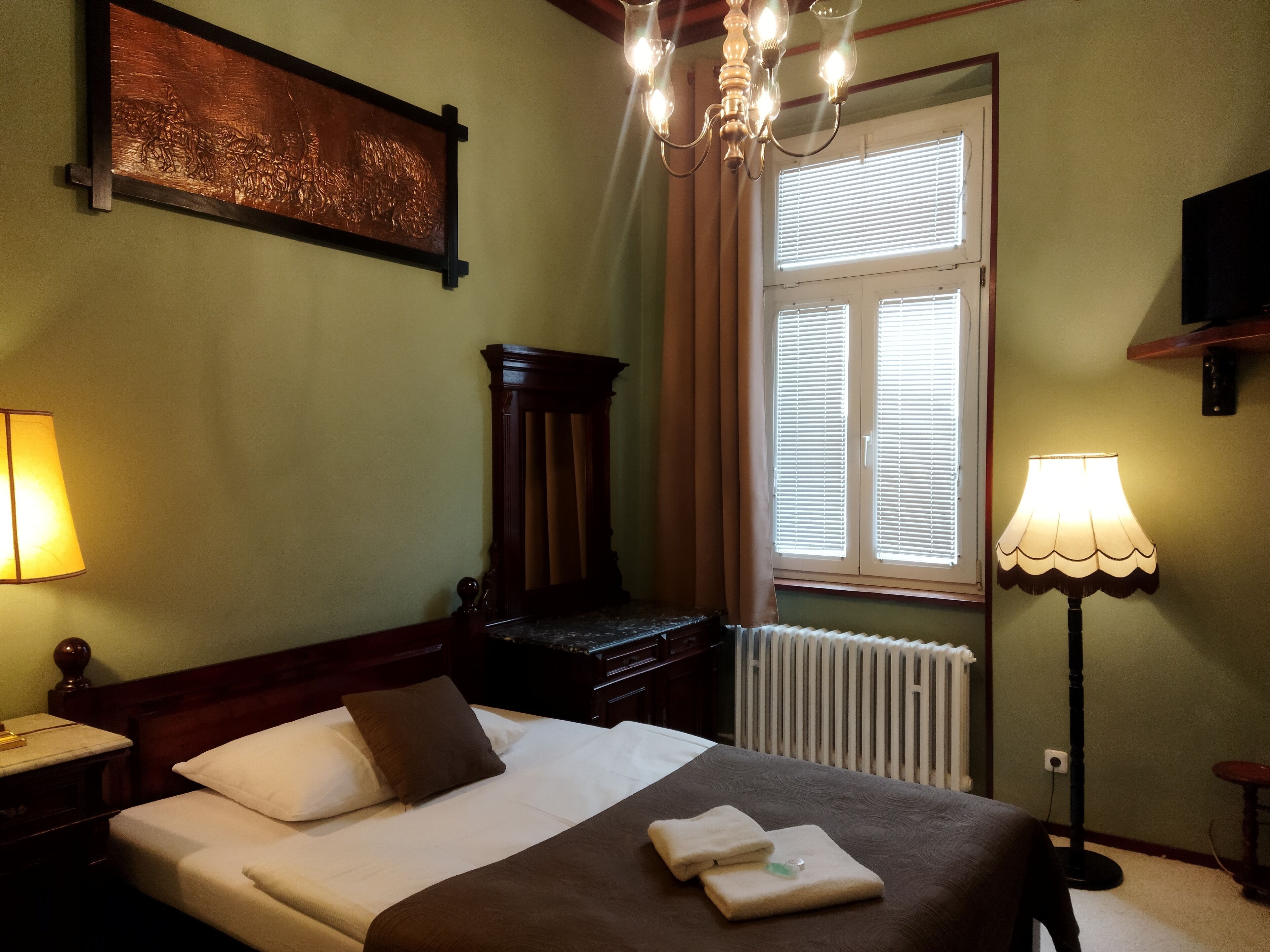 Una camera d'albergo con un letto, un comodino, una lampada, un termosifone e una finestra con persiane.