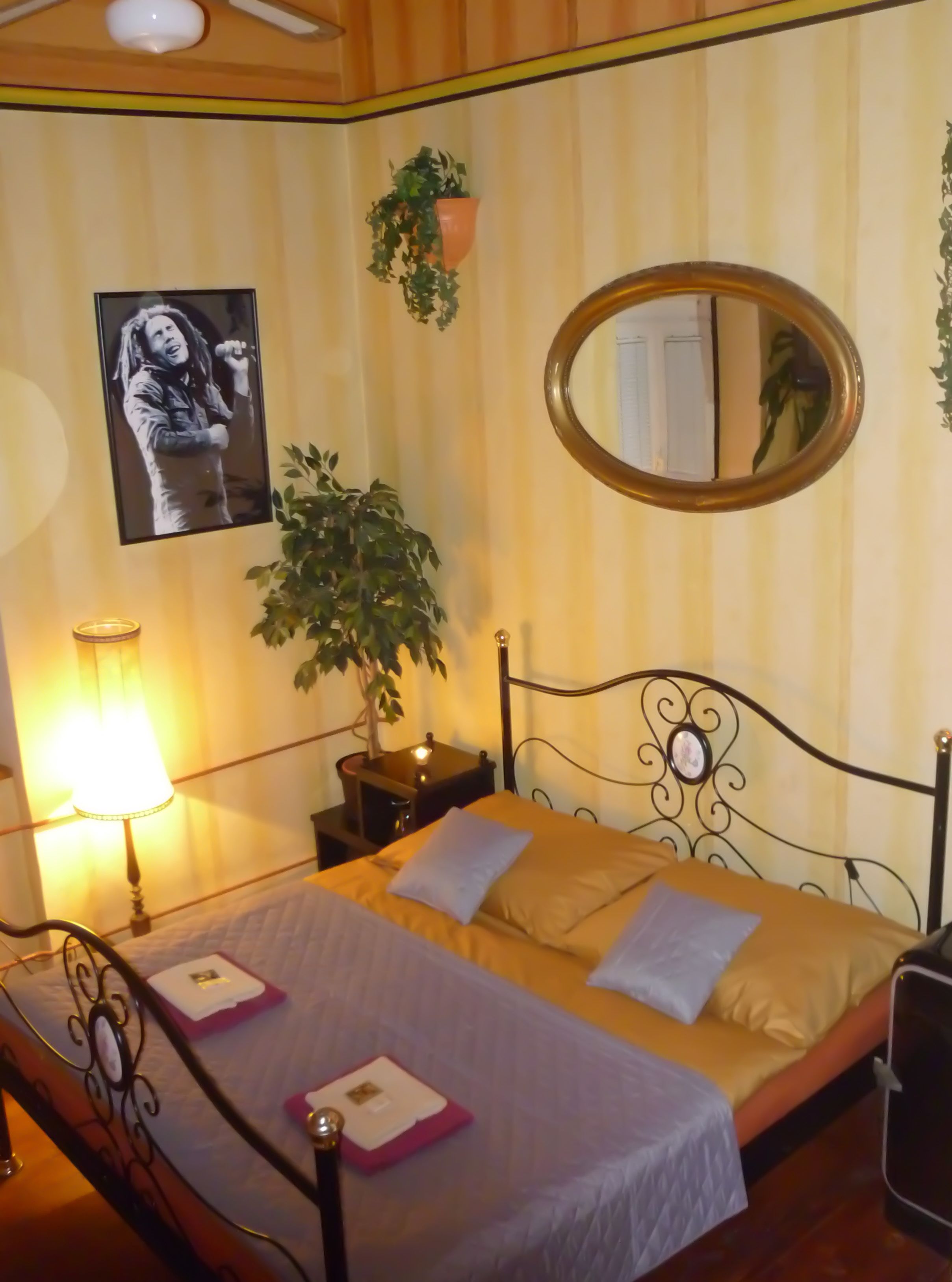 Uma cama de casal com cabeceira, uma mesa de cabeceira com abajur, uma planta em vaso e um quadro na parede.