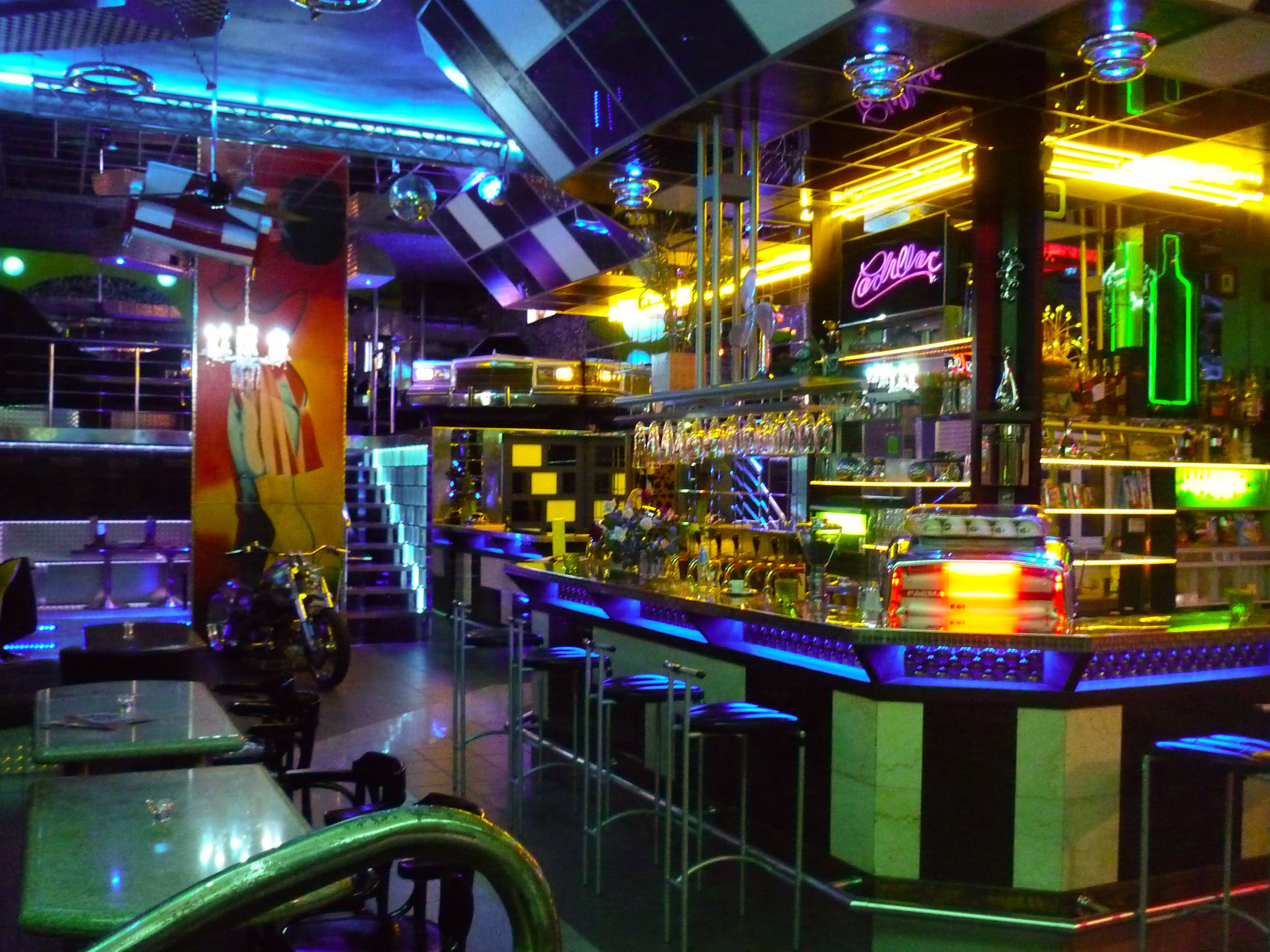 Zona de bar con un letrero de neón de Cadillac, taburetes y una variedad de botellas y vasos en estantes.