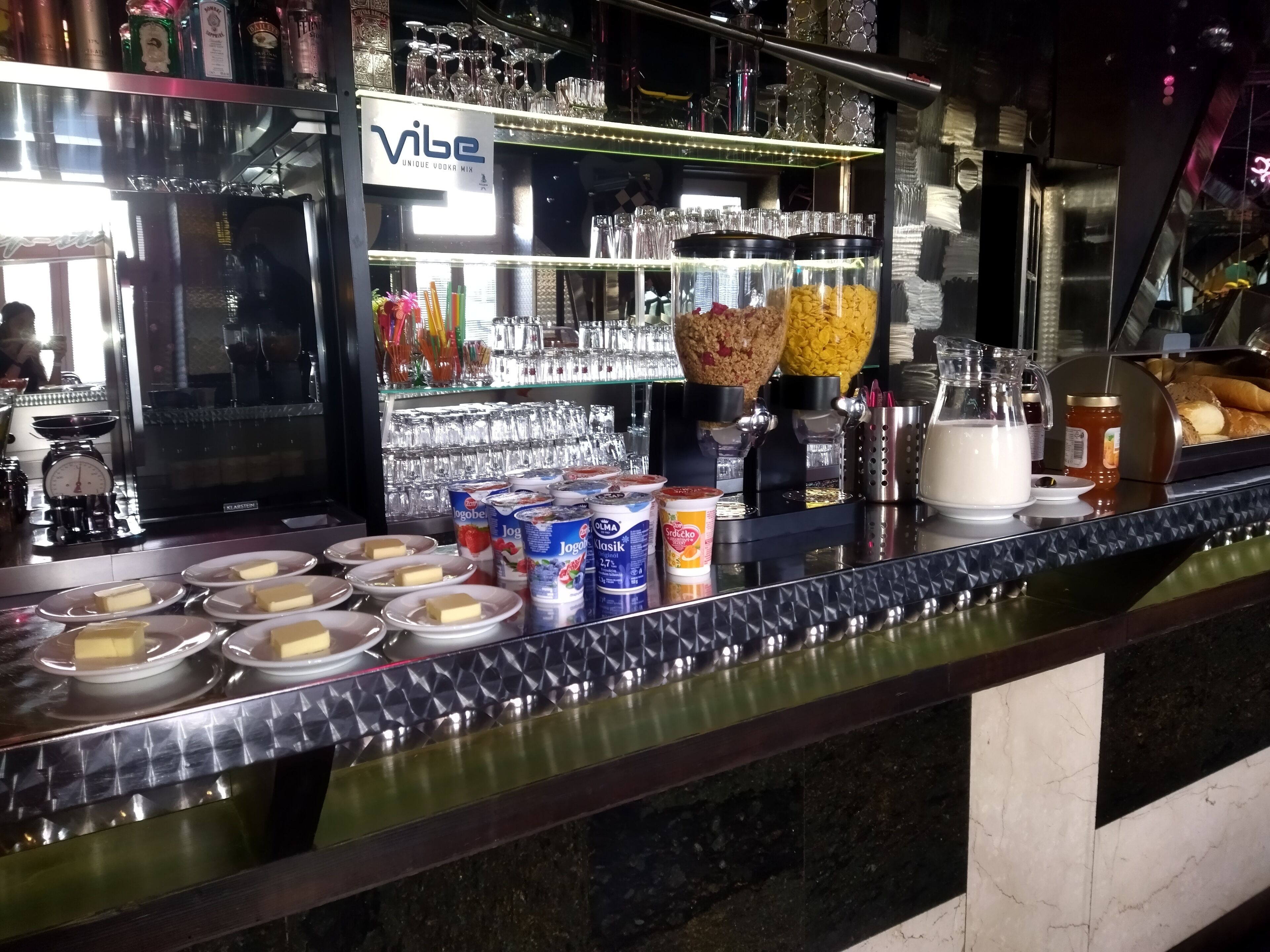 Een bar met een toonbank waarop diverse etenswaren en sauzen staan, een plank met servies en een bordje met de tekst 'Vibe Unique Vodka Mix'.