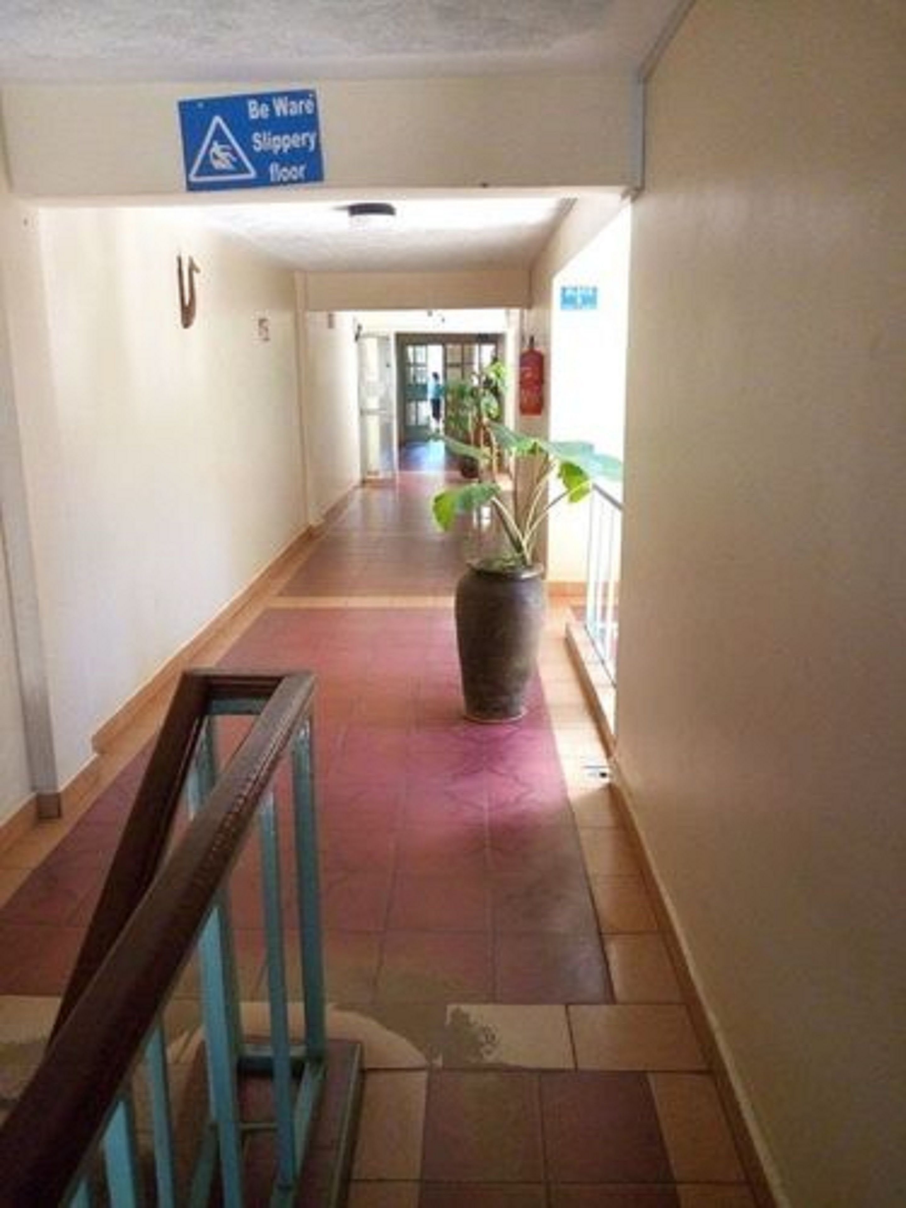 Un pasillo con un cartel de suelo resbaladizo, una planta en maceta y una escalera con barandilla.