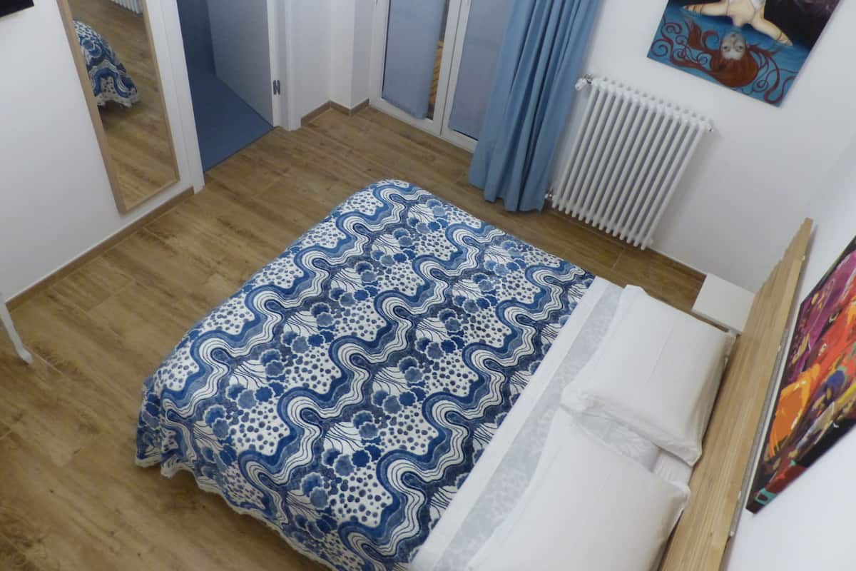 A bedroom with a patterned bedspread, a white mattress, a wooden floor, a radiator, and a painting on the wall.
