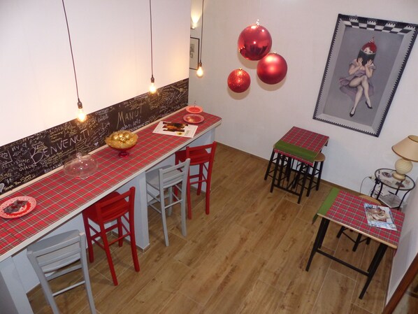 A dining area with a red and white checkered tablecloth, red chairs, and a small table with a green tablecloth. There are three red spherical ornaments on the wall and a framed picture.