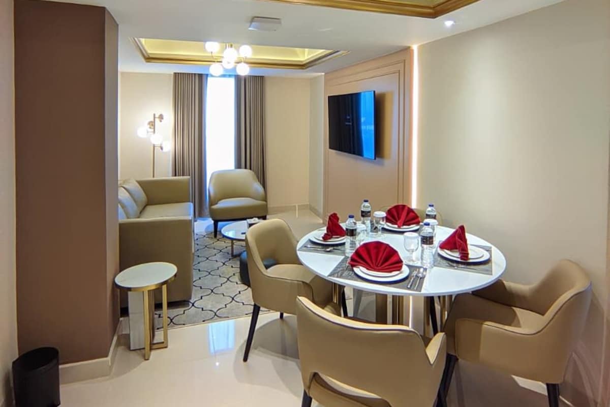 A modern dining area with a round table set for four, a flat-screen TV mounted on the wall, and a ceiling with recessed lighting.