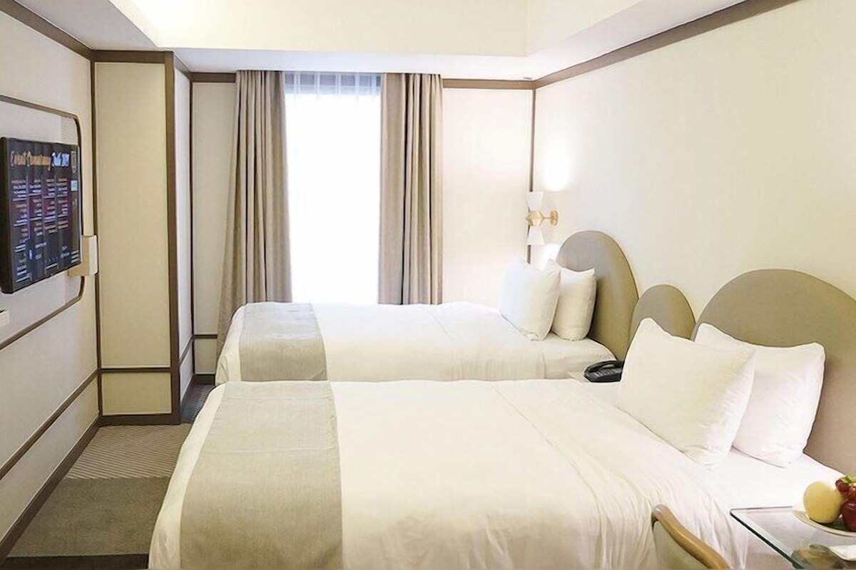 Habitación de hotel con dos camas, un televisor de pantalla plana, un escritorio con lámpara y una ventana con cortinas.