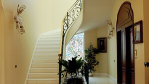 A grand staircase with a black railing, a large potted plant at the bottom, and a stained glass window in the background.