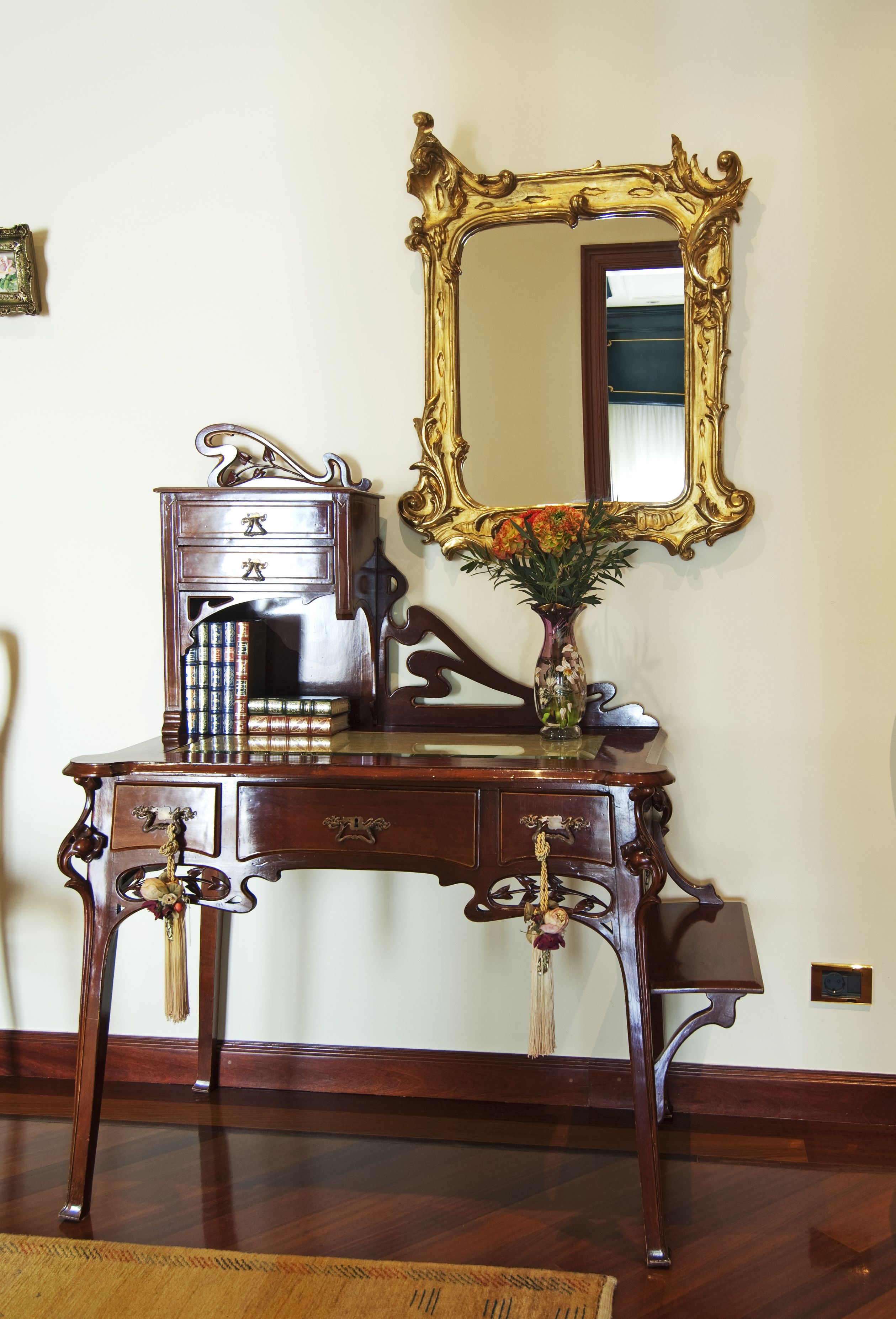 A wooden desk with drawers, a vase with flowers, and a large ornate mirror.