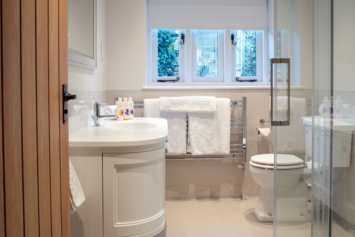 Une salle de bain moderne avec un lavabo blanc, une cabine de douche en verre et des toilettes. Il y a trois fenêtres au-dessus du lavabo, et on peut voir un porte-serviettes.