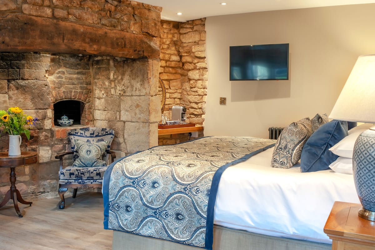 A bedroom with a stone fireplace, a bed with patterned bedding, a chair with a blue and white design, and a flat-screen TV on the wall.