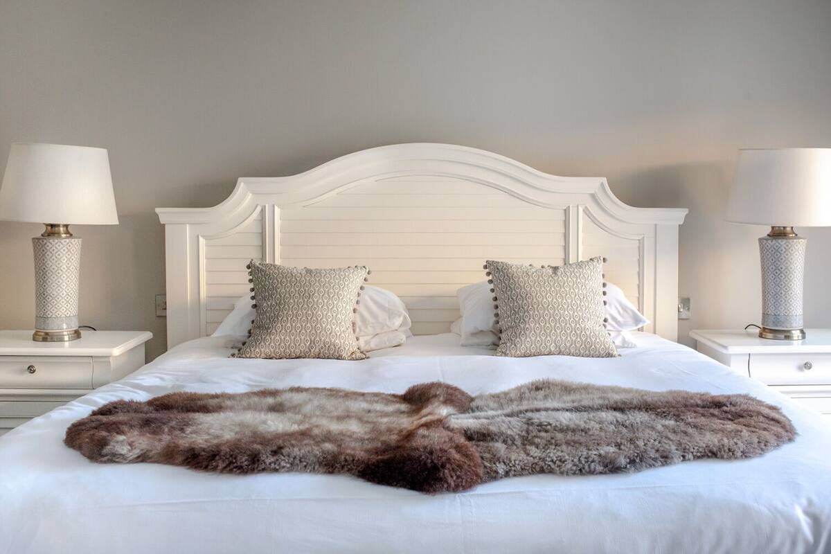 A neatly made bed with a fluffy fur throw, two patterned pillows, and two bedside lamps on white nightstands.