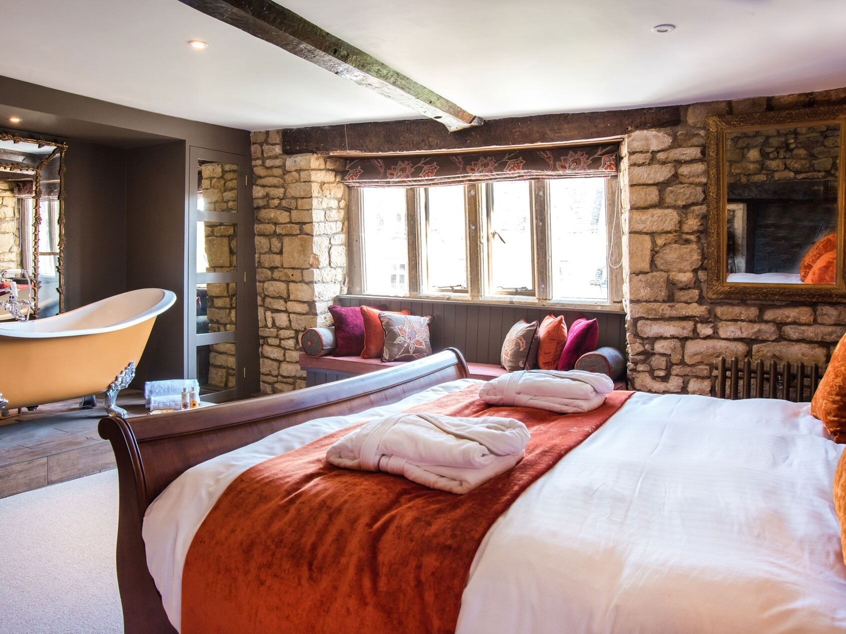 A bedroom with a stone wall, a large bed with a red blanket, a wooden bench with pillows, a bathtub, and a fireplace.