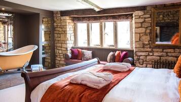 A bedroom with a stone wall, a large bed with a red blanket, a wooden bench with pillows, a bathtub, and a fireplace.