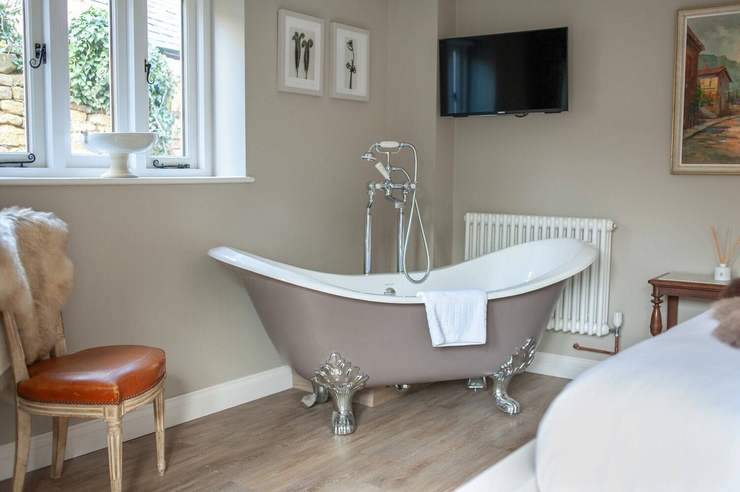 A bathroom with a freestanding bathtub, a wooden chair, a radiator, and a television mounted on the wall.