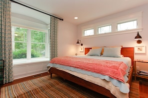 A bedroom with a large bed, a window with curtains, a bedside table with a lamp, and a view of greenery outside. - Farmhouse (Saugatuck)