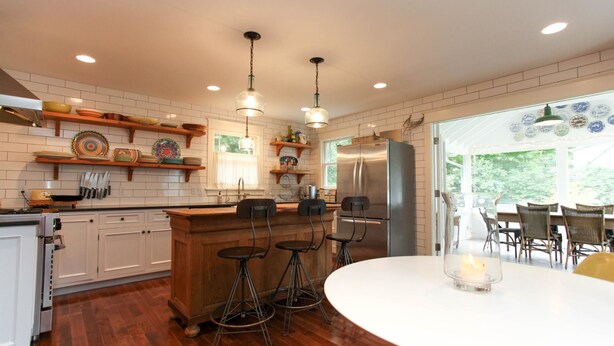 A modern kitchen with white cabinets, wooden countertops, and a central island with stools. There is a large refrigerator and a dining area with a table and chairs. - Farmhouse (Saugatuck)