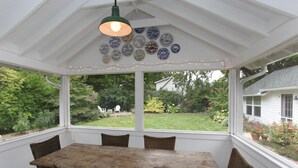 A covered patio with a wooden table and wicker chairs, a hanging light fixture, and a view of a garden and house. - Farmhouse (Saugatuck)