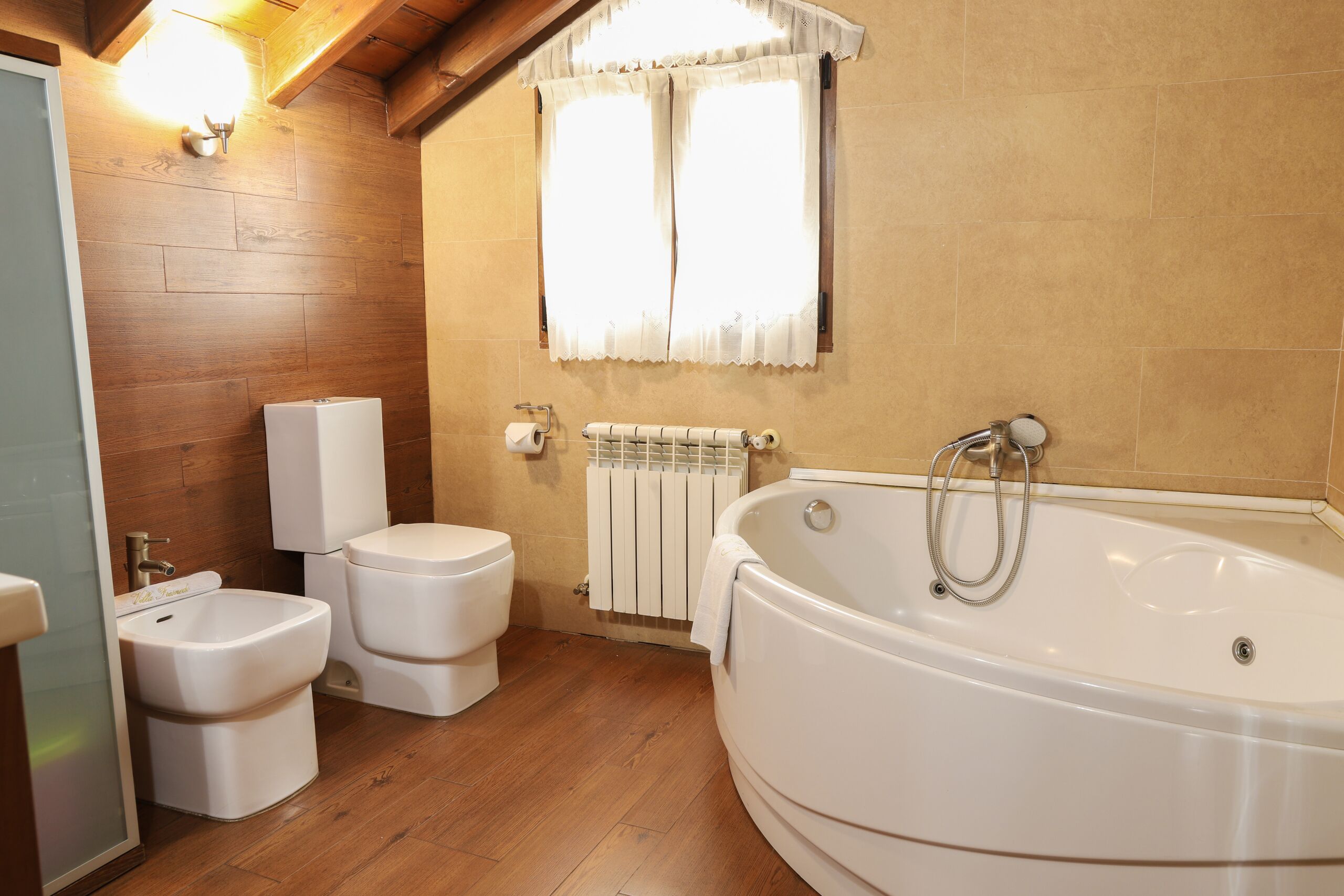 Une salle de bain avec un plancher en bois, une baignoire, deux toilettes et une fenêtre avec des rideaux.