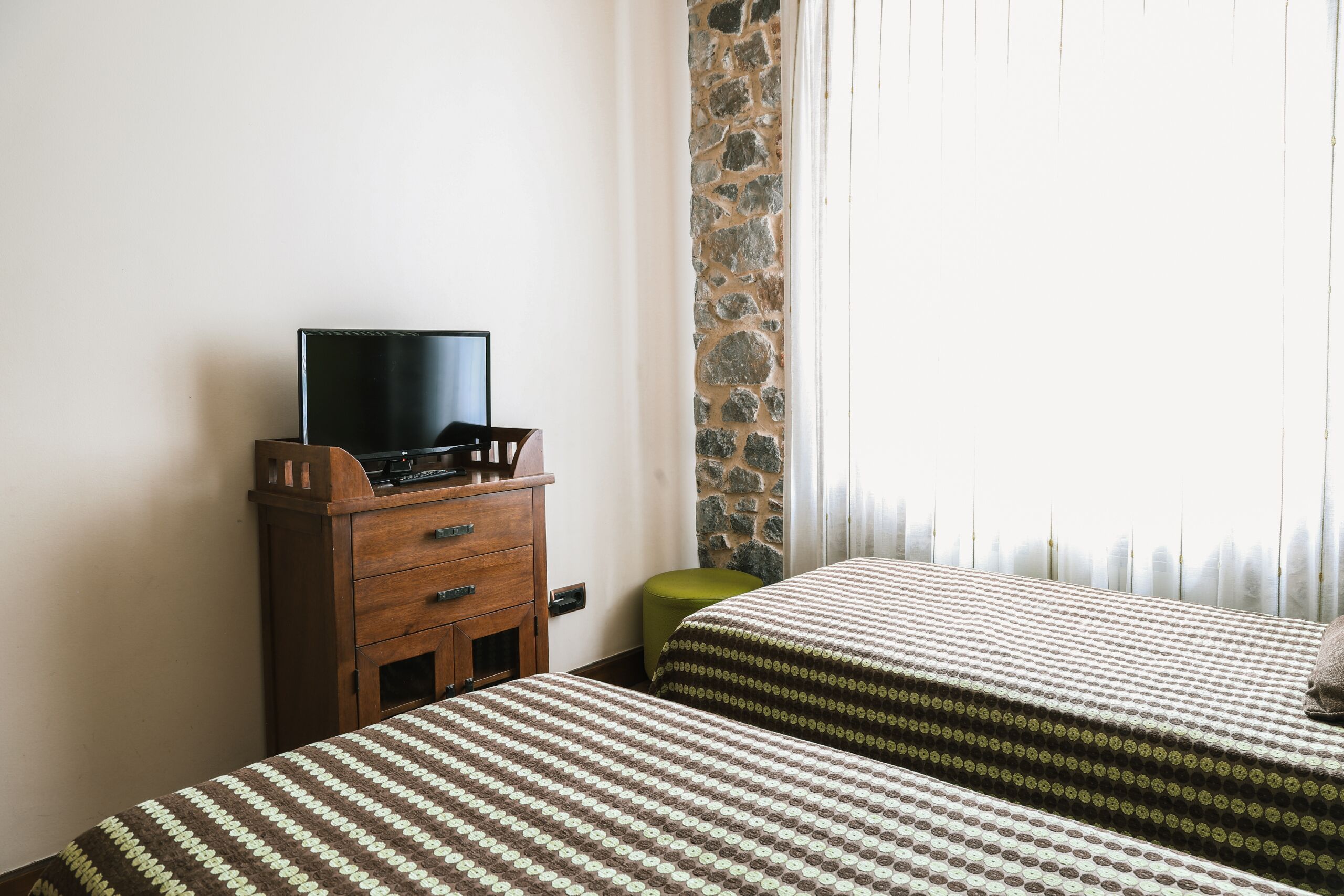 A hotel room with two beds, a wooden dresser, a television on the dresser, and a stone wall.